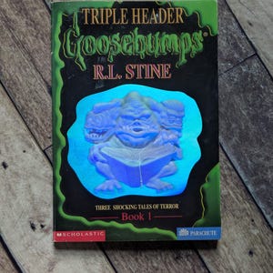 以下が含まれることがあります： R.L.スタイン著の「Triple Header」というタイトルのヴィンテージ「Goosebumps」の本。表紙には、本を読む三つ頭のモンスターが描かれており、「Three Shocking Tales of Terror」と「Book 1」というテキストがあります。本は木製の表面にあります。