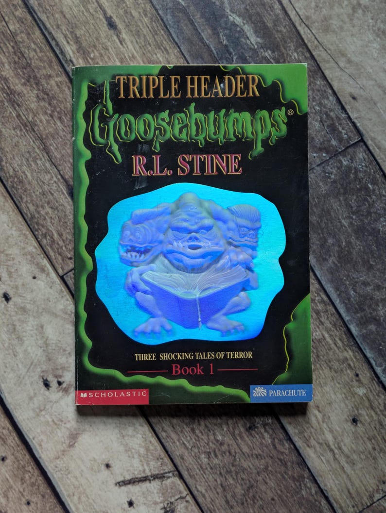 以下が含まれることがあります： R.L.スタイン著の「Triple Header」というタイトルのヴィンテージ「Goosebumps」の本。表紙には、本を読む三つ頭のモンスターが描かれており、「Three Shocking Tales of Terror」と「Book 1」というテキストがあります。本は木製の表面にあります。