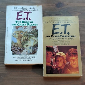 May include: Two vintage paperback books, "E.T. The Book of the Green Planet" and "E.T. The Extra-Terrestrial." The first book features E.T. on the cover, while the second shows children. Both books are on a wooden surface.