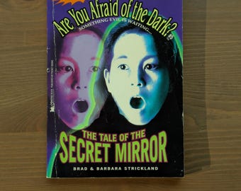 Puede incluir: Un libro de bolsillo titulado "Are You Afraid of the Dark? The Tale of the Secret Mirror" de Brad & Barbara Strickland. La portada presenta dos caras con la boca abierta, un fondo morado y el logotipo de Nickelodeon.