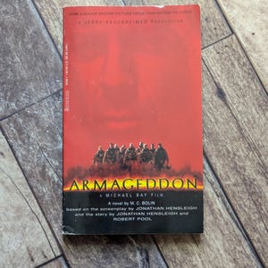 Könnte beinhalten: Ein Taschenbuch mit dem Titel "Armageddon" mit rotem Einband. Auf dem Cover sind eine Gruppe von Silhouetten und der Filmtitel abgebildet. Das Buch ist ein Roman von M.C. Bolin.