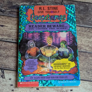 May include: A paperback book titled Give Yourself Goosebumps #19 Escape From Camp Run-For-Your-Life by R.L. Stine. The cover features a holographic foil background with an illustration of three monsters around a campfire and text.