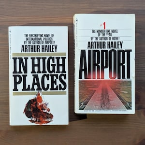 May include: Two vintage books by Arthur Hailey are displayed on a wooden surface. The book on the left is titled "In High Places" and the book on the right is titled "Airport". Both books have white covers with black and red text.