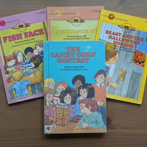 May include: Four vintage children's books with colorful covers are arranged on a wooden surface. Titles include "Fish Face," "Sunny-Side Up," "The Candy Corn Contest," and "Beast and the Halloween Horror." The books feature illustrations and text.