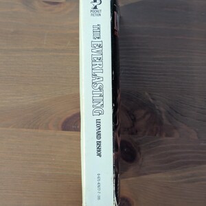 May include: A paperback book titled "The Everlasting" by Leonard Bishop. The spine is white with black text, featuring the Pocket Fiction logo. The book is set against a wooden background.
