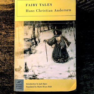 May include: A paperback book titled Fairy Tales by Hans Christian Andersen, published by Barnes & Noble Classics. The cover features a yellow top and bottom border with a central illustration of a person in a dark coat walking in snow.
