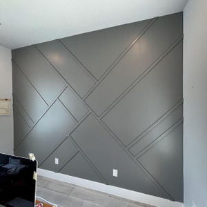 Op de afbeelding: Een grijze accentmuur met een geometrisch patroon gecreëerd door het toevoegen van houten panelen. De muur heeft een modern ontwerp en is geverfd in een lichtgrijs.