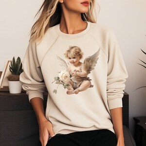 May include: Beige sweatshirt featuring a detailed illustration of a cherub holding a white rose. The cherub has wings and is seated. The sweatshirt has long sleeves and a crew neck.