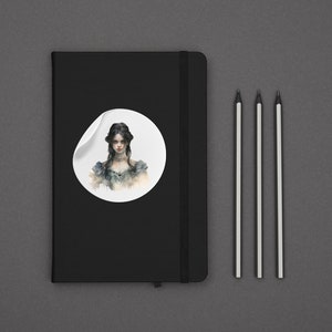 K&ouml;nnte beinhalten: Ein schwarzes Notizbuch mit einem wei&szlig;en Aufkleber, der eine Aquarell-Illustration einer Frau mit dunklen Haaren und einem wei&szlig;en Kleid zeigt. Der Aufkleber befindet sich auf dem vorderen Einband des Notizbuchs. Drei Bleistifte liegen auf einer grauen Oberfl&auml;che.