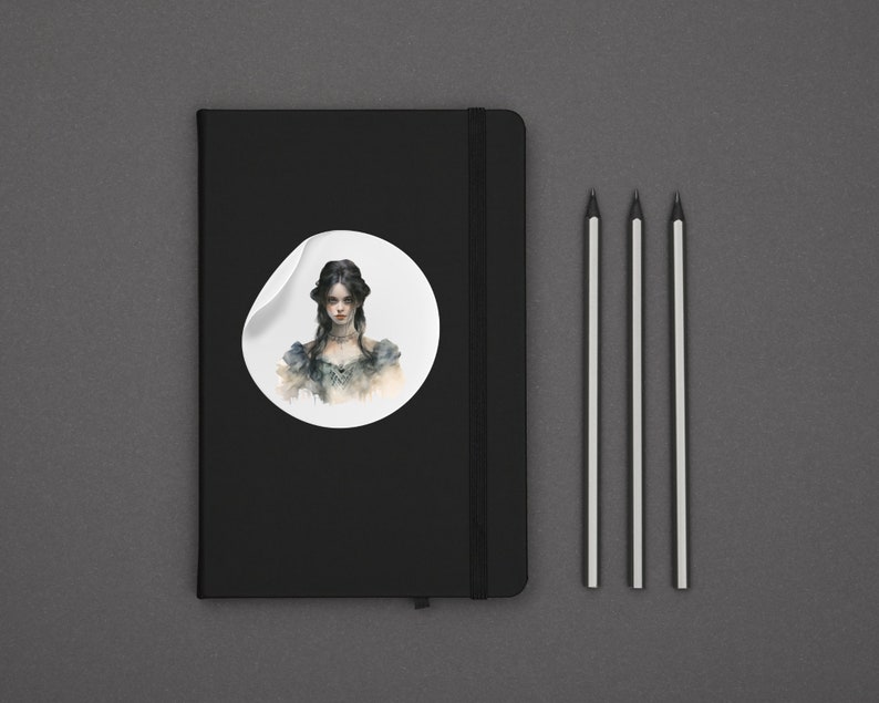 K&ouml;nnte beinhalten: Ein schwarzes Notizbuch mit einem wei&szlig;en Aufkleber, der eine Aquarell-Illustration einer Frau mit dunklen Haaren und einem wei&szlig;en Kleid zeigt. Der Aufkleber befindet sich auf dem vorderen Einband des Notizbuchs. Drei Bleistifte liegen auf einer grauen Oberfl&auml;che.