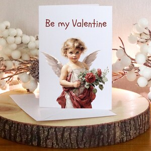 May include: A Valentine's Day greeting card featuring a vintage illustration of a cherub holding roses, with the text "Be my Valentine" in red. The card is displayed on a wooden slice, with a white envelope and decorative white berries in the background.