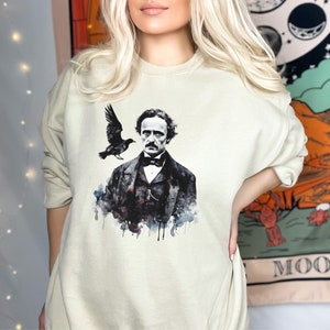 K&ouml;nnte beinhalten: Ein hellbeiger Pullover mit einer Aquarell-Illustration von Edgar Allan Poe und einem Raben. Die Illustration ist in Schwarz, Wei&szlig; und Grau.