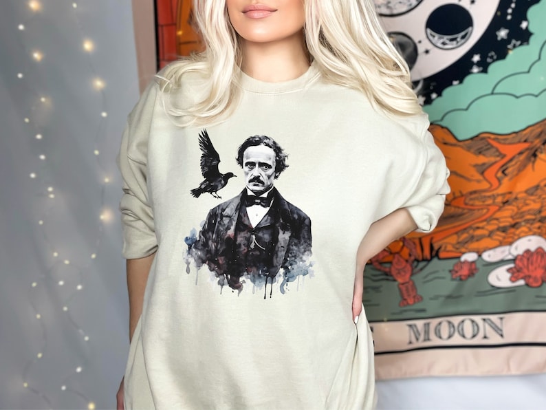 K&ouml;nnte beinhalten: Ein hellbeiger Pullover mit einer Aquarell-Illustration von Edgar Allan Poe und einem Raben. Die Illustration ist in Schwarz, Wei&szlig; und Grau.