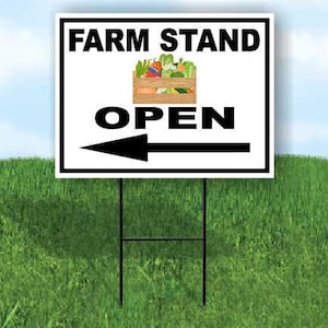 May include: A white sign with black text that reads "FARM STAND OPEN" with an arrow pointing left. The sign has a graphic of a wooden crate filled with colorful vegetables. The sign is on a black metal stand, set in a grassy field.