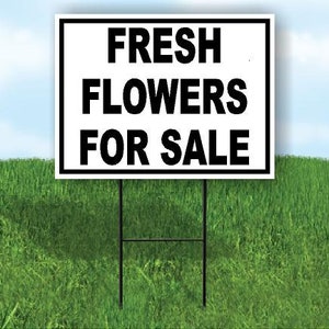 May include: A white sign with black text that reads "FRESH FLOWERS FOR SALE" is displayed on a black metal stake in a grassy field.