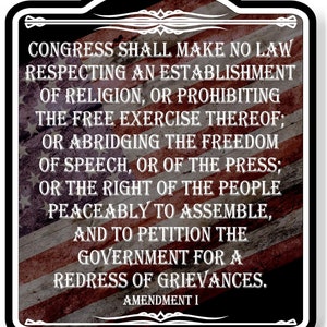 May include: A black and white sign with a red, white, and blue American flag background. The sign features the text of the First Amendment to the United States Constitution: "Congress shall make no law respecting an establishment of religion, or prohibiting the free exercise thereof; or abridging the freedom of speech, or of the press; or the right of the people peaceably to assemble, and to petition the government for a redress of grievances." The sign also includes the text "Amendment I".
