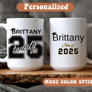 May include: Two white ceramic mugs with black text and a graduation cap graphic. One mug says "Brittany 25 twenty" and the other says "Brittany Class of 2025". The mugs are on a wooden surface with the text "Personalized" and "More Color Options".