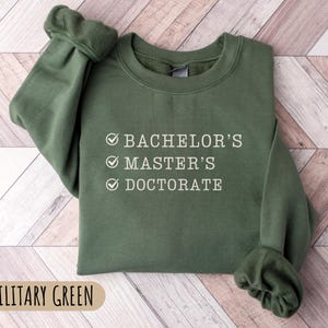 May include: A green sweatshirt with the text "Bachelor's, Master's, Doctorate" checked off in white. The sweatshirt is on a woodgrain background.