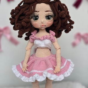 May include: A handmade amigurumi doll with brown curly hair, wearing a pink and white dress. The doll has a detailed face with large eyes and long eyelashes. The doll is standing on a wooden base with the text "ilmekilmekbebek".