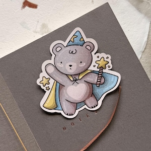 A charming cottagecore die-cut sticker featuring a teddy bear dressed as a wizard, complete with a pointed hat, flowing cape, and a magic wand sparkling with stars. Perfect for adding a touch of whimsy to laptops, water bottles, and journals.