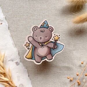 A charming cottagecore die-cut sticker featuring a teddy bear dressed as a wizard, complete with a pointed hat, flowing cape, and a magic wand sparkling with stars. Perfect for adding a touch of whimsy to laptops, water bottles, and journals.