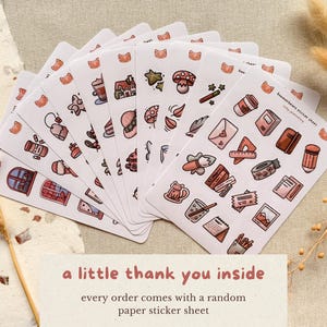 Flat lay photo of multiple small paper sticker sheets fanned out on a neutral linen background, featuring hand-drawn cozy journaling illustrations in warm pink and brown tones. Dried wheat and paper textures surround the stickers. Text on the image reads: “a little thank you inside – every order comes with a random paper sticker sheet.”
