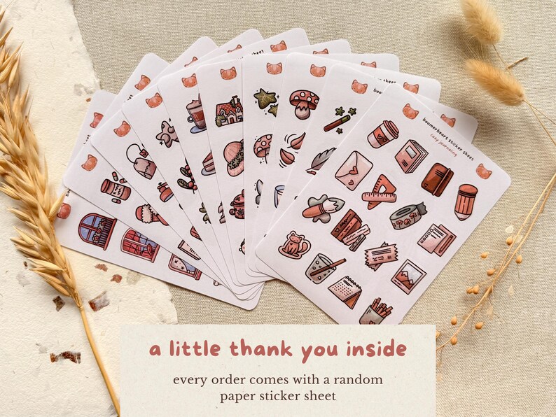 Flat lay photo of multiple small paper sticker sheets fanned out on a neutral linen background, featuring hand-drawn cozy journaling illustrations in warm pink and brown tones. Dried wheat and paper textures surround the stickers. Text on the image reads: “a little thank you inside – every order comes with a random paper sticker sheet.”