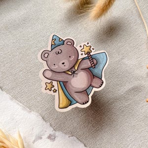 A charming cottagecore die-cut sticker featuring a teddy bear dressed as a wizard, complete with a pointed hat, flowing cape, and a magic wand sparkling with stars. Perfect for adding a touch of whimsy to laptops, water bottles, and journals.