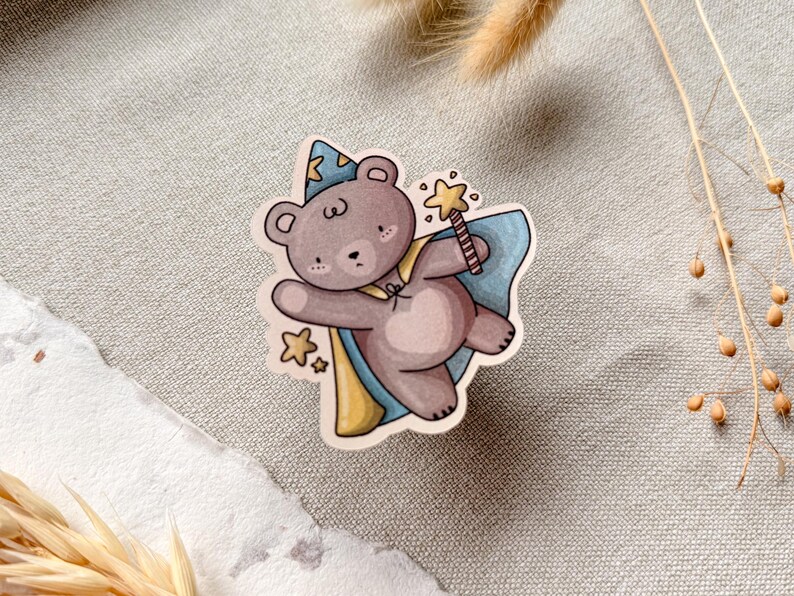 A charming cottagecore die-cut sticker featuring a teddy bear dressed as a wizard, complete with a pointed hat, flowing cape, and a magic wand sparkling with stars. Perfect for adding a touch of whimsy to laptops, water bottles, and journals.