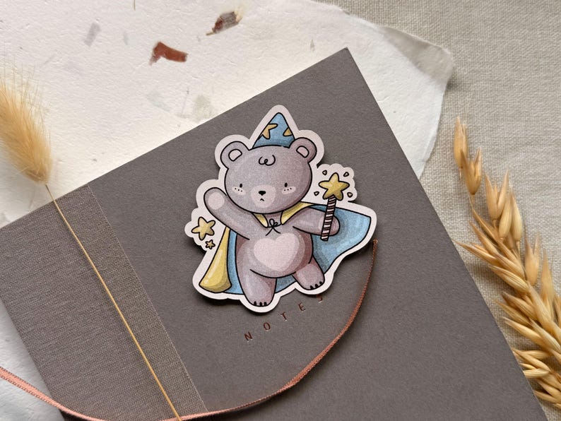 A charming cottagecore die-cut sticker featuring a teddy bear dressed as a wizard, complete with a pointed hat, flowing cape, and a magic wand sparkling with stars. Perfect for adding a touch of whimsy to laptops, water bottles, and journals.
