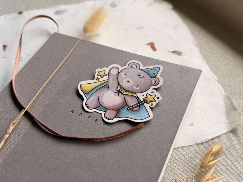 A charming cottagecore die-cut sticker featuring a teddy bear dressed as a wizard, complete with a pointed hat, flowing cape, and a magic wand sparkling with stars. Perfect for adding a touch of whimsy to laptops, water bottles, and journals.