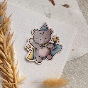 A charming cottagecore die-cut sticker featuring a teddy bear dressed as a wizard, complete with a pointed hat, flowing cape, and a magic wand sparkling with stars. Perfect for adding a touch of whimsy to laptops, water bottles, and journals.