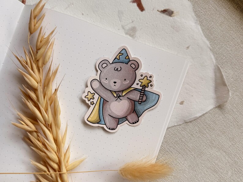 A charming cottagecore die-cut sticker featuring a teddy bear dressed as a wizard, complete with a pointed hat, flowing cape, and a magic wand sparkling with stars. Perfect for adding a touch of whimsy to laptops, water bottles, and journals.