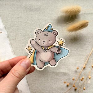 A charming cottagecore die-cut sticker featuring a teddy bear dressed as a wizard, complete with a pointed hat, flowing cape, and a magic wand sparkling with stars. Perfect for adding a touch of whimsy to laptops, water bottles, and journals.