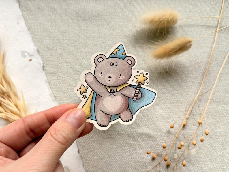 A charming cottagecore die-cut sticker featuring a teddy bear dressed as a wizard, complete with a pointed hat, flowing cape, and a magic wand sparkling with stars. Perfect for adding a touch of whimsy to laptops, water bottles, and journals.
