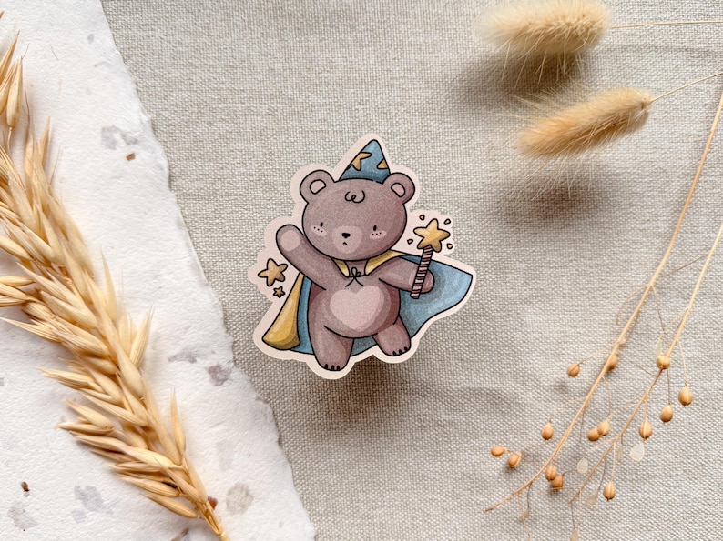 A charming cottagecore die-cut sticker featuring a teddy bear dressed as a wizard, complete with a pointed hat, flowing cape, and a magic wand sparkling with stars. Perfect for adding a touch of whimsy to laptops, water bottles, and journals.