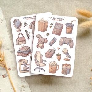 Könnte beinhalten: Zwei Stickerbögen mit Illustrationen von gemütlichen Gegenständen. Ein Bogen zeigt einen Spiegel, eine Handtasche und eine Kaffeemaschine mit dem Text "Messy Girl Mornings". Der andere Bogen hat eine Spielkonsole, einen Controller und Kaffee mit dem Text "Cozy Gamer Comforts."