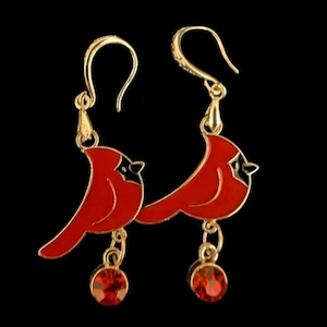 Puede incluir: Pendientes dorados con diseños de pájaros cardenales rojos y detalles en negro. Cada pendiente tiene una gema roja colgante. Fondo negro.