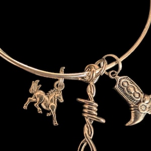 May include: Gold-toned bracelet with three charms: a horse, a barbed wire design, and a cowboy boot. The bracelet is set against a black background, highlighting the metallic sheen of the charms.