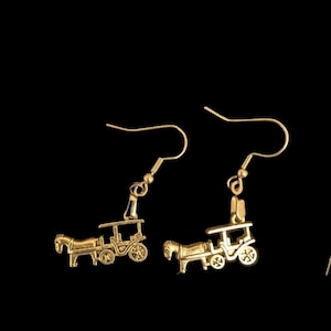 May include: A pair of gold-colored earrings featuring a horse-drawn carriage design. The earrings have a hook closure and are set against a black background. The carriage has detailed wheels and a roof.