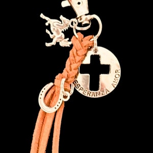 May include: A silver-toned keychain with a horse charm, a horseshoe charm, and a circular charm with a cross cutout. The circular charm has the words "AMOR" and "ESPERANZA" engraved on it. The keychain has a braided tan leather strap.
