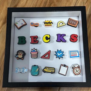 May include: A black frame with a white background displaying colorful school-themed magnets. The magnets include a book, pencils, a protractor, a chalkboard, letters spelling "BECKS", a penguin, a pencil holder, a triangle, a check mark, an abacus, a backpack, a school bus, a clipboard, and an alarm clock.