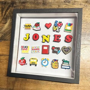 May include: A black framed shadow box with a white background and various colorful school-themed decorations. The decorations include a red heart, a yellow school bus, a red apple, a red notebook, a yellow and red crayon box, and a yellow and red alphabet block. The name "JONES" is spelled out in large, colorful letters.