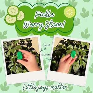 May include: A green, textured pickle-shaped worry stone. The image features two polaroid-style photos of the object held against a leafy background, with the text Pickle Worry Stone! at the top and Little joys matter. at the bottom of the graphic.