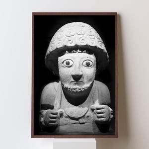 Pode incluir: Fotografia a preto e branco de uma escultura em pedra de uma figura com olhos grandes, barba e um cocar padronizado. A escultura está emoldurada numa moldura de madeira escura e montada num pedestal branco.