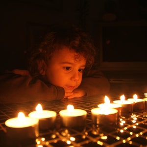 Pode incluir: Uma crian&ccedil;a pequena com cabelo castanho encaracolado senta-se em um quarto escuro, olhando para uma fileira de velas acesas em uma superf&iacute;cie de azulejos. As velas est&atilde;o em pequenos porta-velas de vidro e emitem um brilho quente.