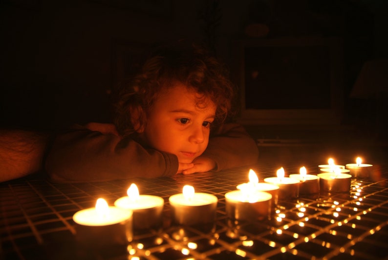Pode incluir: Uma crian&ccedil;a pequena com cabelo castanho encaracolado senta-se em um quarto escuro, olhando para uma fileira de velas acesas em uma superf&iacute;cie de azulejos. As velas est&atilde;o em pequenos porta-velas de vidro e emitem um brilho quente.