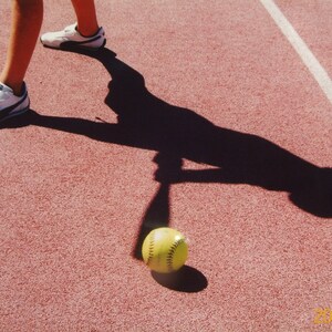 Pode incluir: Uma pessoa com t&ecirc;nis brancos corre em uma pista vermelha com uma sombra dela mesma balan&ccedil;ando um taco e uma bola de softball amarela no ch&atilde;o.