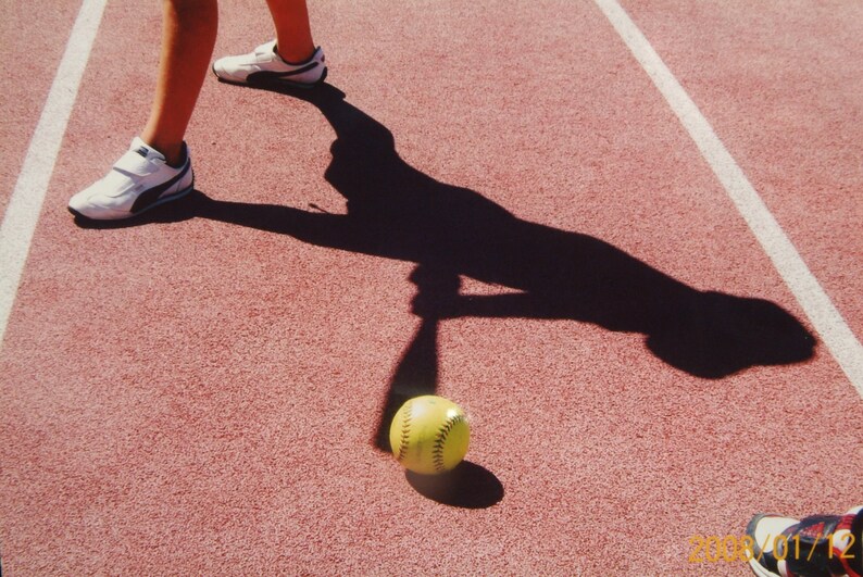 Pode incluir: Uma pessoa com t&ecirc;nis brancos corre em uma pista vermelha com uma sombra dela mesma balan&ccedil;ando um taco e uma bola de softball amarela no ch&atilde;o.
