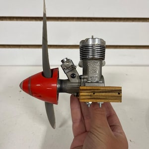 May include: A model airplane engine with a red nose cone and a gray propeller. The engine is silver with the text "ENYA 29 MODEL 5224" printed on the side. The engine is mounted on a wooden base.